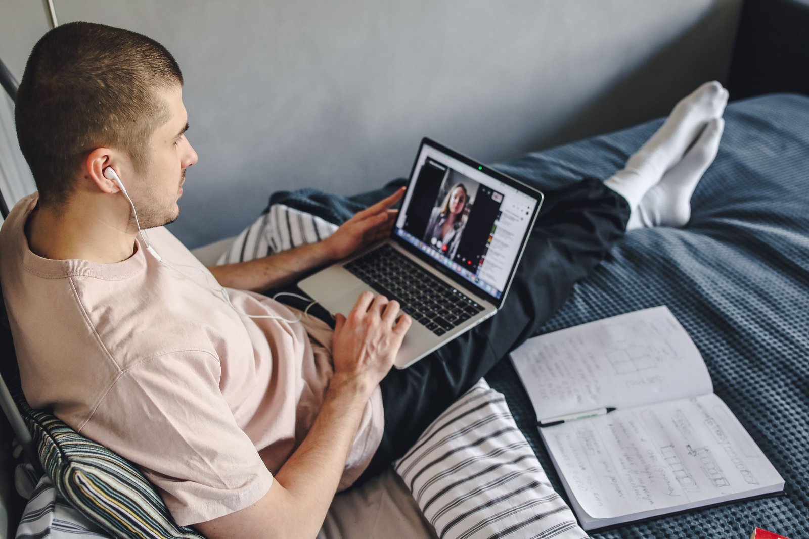 Young man on bed video calling using laptop and earphones, surrounded by notes.